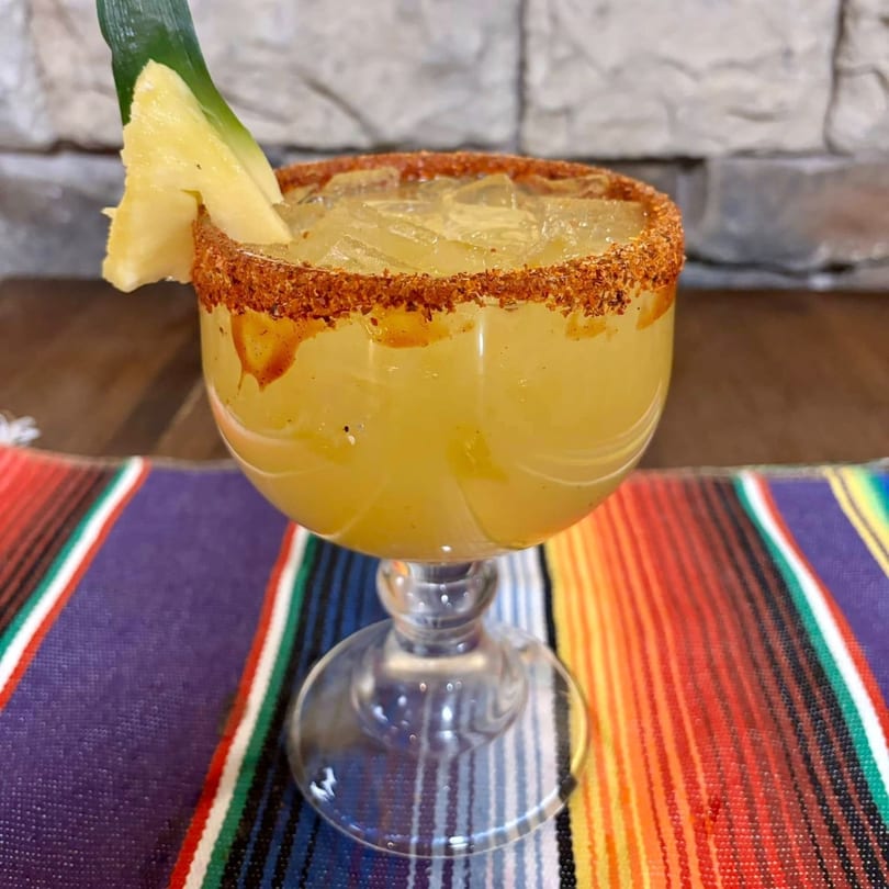 A pineapple margarita with a chili-lime rim and a pineapple wedge, on a colorful striped placemat.