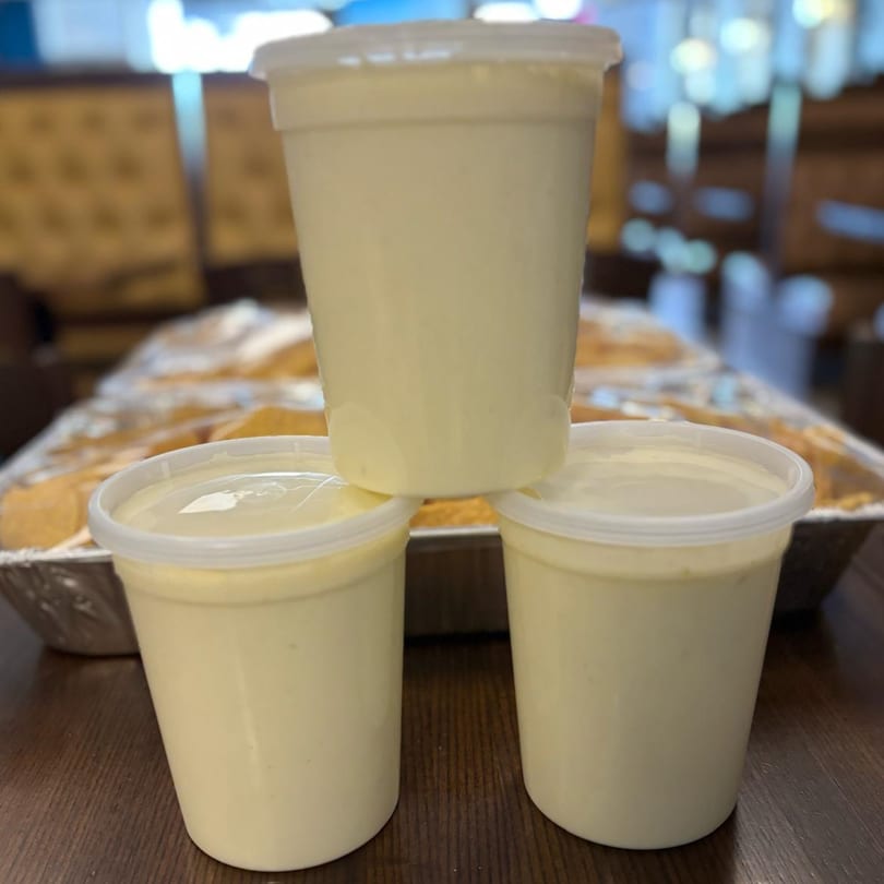 Three plastic containers of El Sazon Mexican Grill's creamy white sauce, with a tray of food in the background.