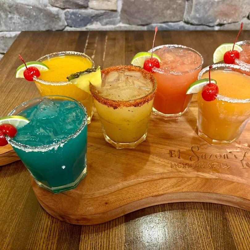 Colorful margaritas with fruit garnishes on a wooden serving board with the El Sazon Mexican Grill logo.