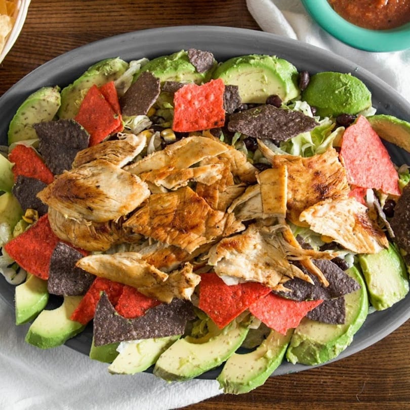 Grilled chicken salad with avocado, red and blue tortilla chips, and lettuce on a gray plate.