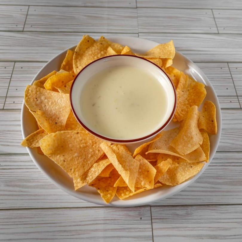 A white bowl of creamy queso dip surrounded by golden tortilla chips on a white plate.