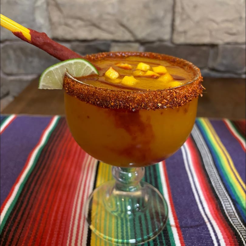 A mango margarita with chili-lime rim, lime wedge, and tamarind candy straw on a colorful striped placemat.