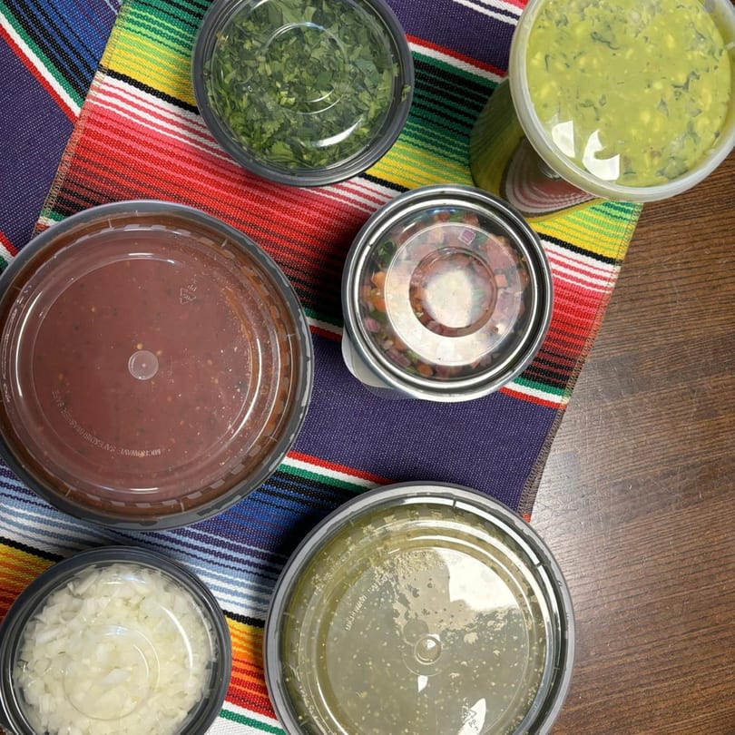 Top-down view of six plastic containers filled with Mexican condiments on a colorful striped serape.