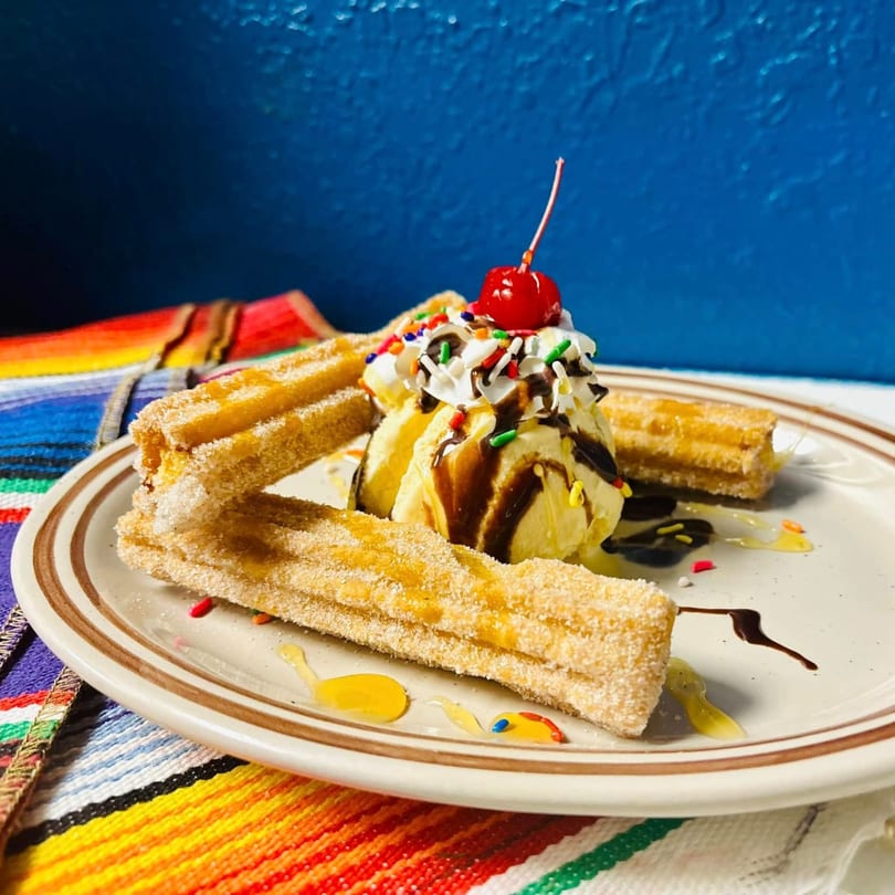 Churros with vanilla ice cream, whipped cream, sprinkles, chocolate, and a cherry on a colorful placemat.