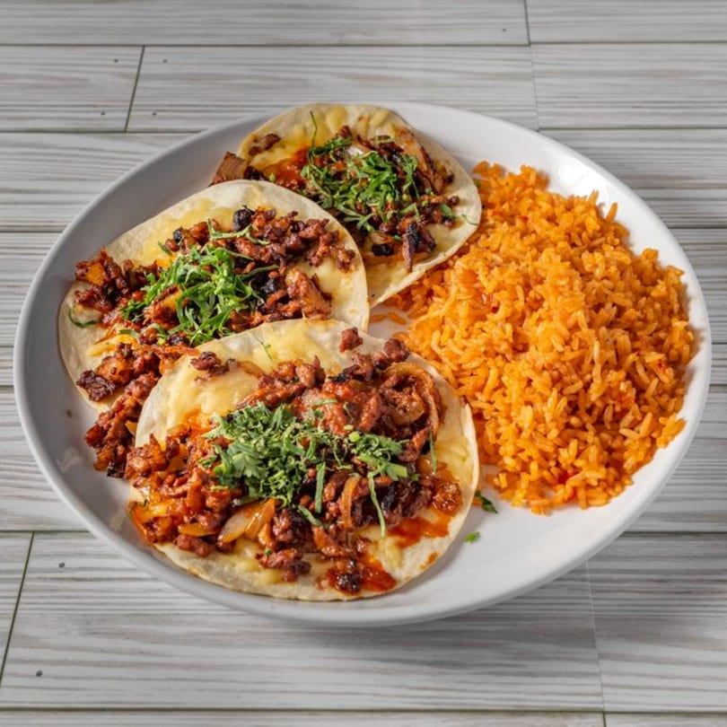 Three El Sazon Mexican Grill tacos with meat, cilantro, and rice on a white plate.
