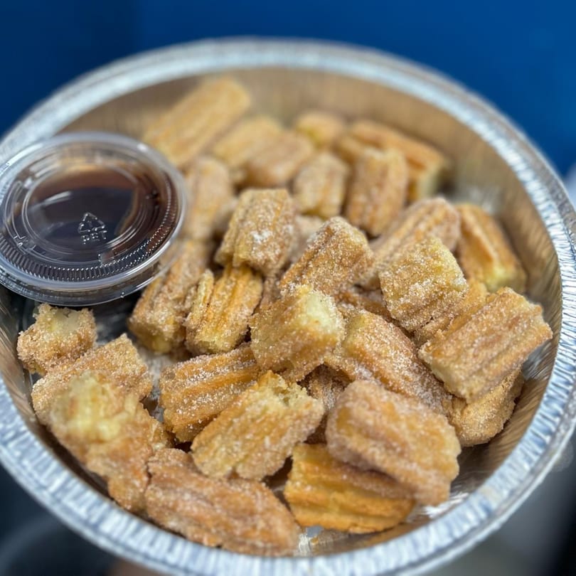 A foil container filled with mini churros dusted with cinnamon sugar, alongside a small cup of dipping sauce.