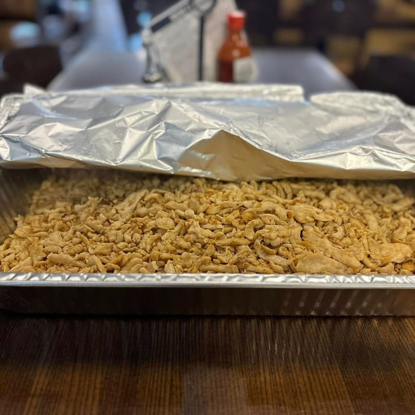 A large aluminum tray filled with seasoned shredded chicken, partially covered with foil, on a wooden table.