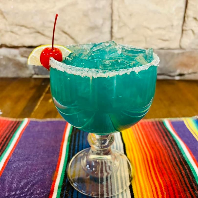 Blue margarita with a salted rim, lemon slice, and cherry on a colorful striped placemat.