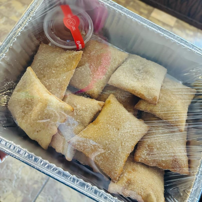 A foil container filled with golden-brown, sugar-dusted Mexican dessert empanadas and a small sauce cup.