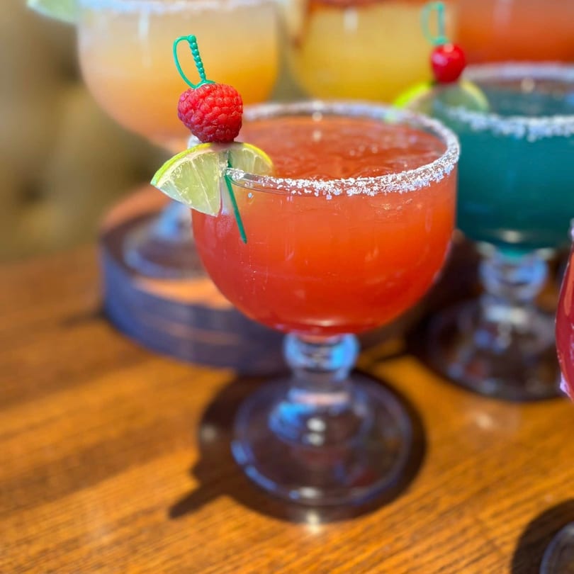 A vibrant red margarita with a salted rim, garnished with a lime wedge and raspberry, on a wooden table.