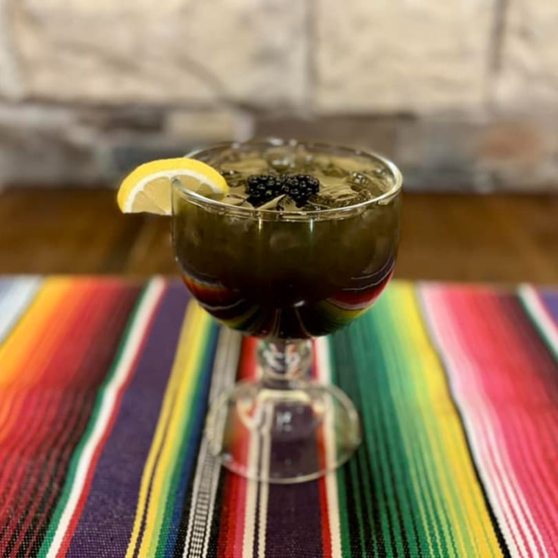 Dark beverage with ice, blackberries, and a lemon slice in a goblet on a colorful striped serape.